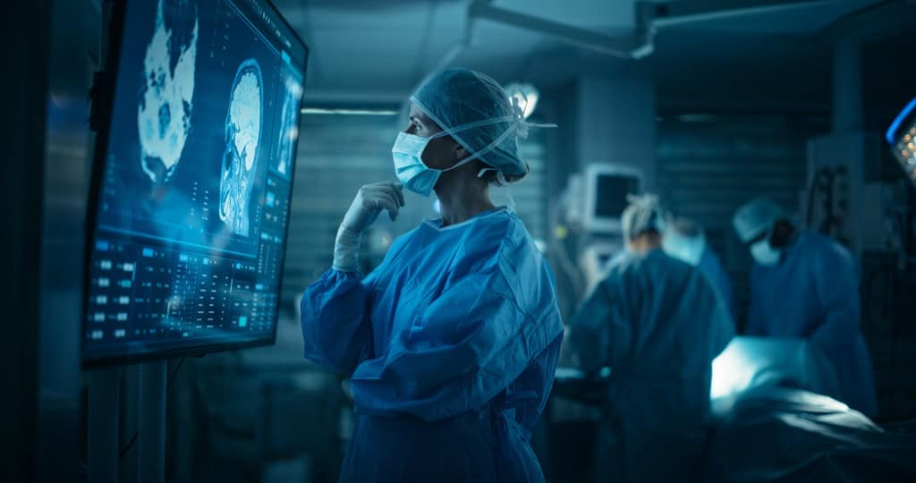 Cirurgiã paramentada com roupas cirúrgicas com a mão no queixo e olhando uma tomografia computadorizada de uma cabeça humana