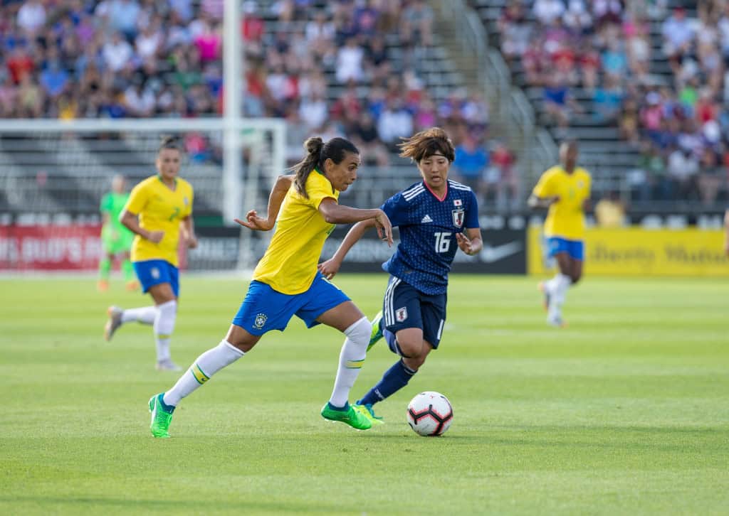Jogadora de futebol Marta carregando a bola e sendo marcada de perto por uma jogadora japonesa