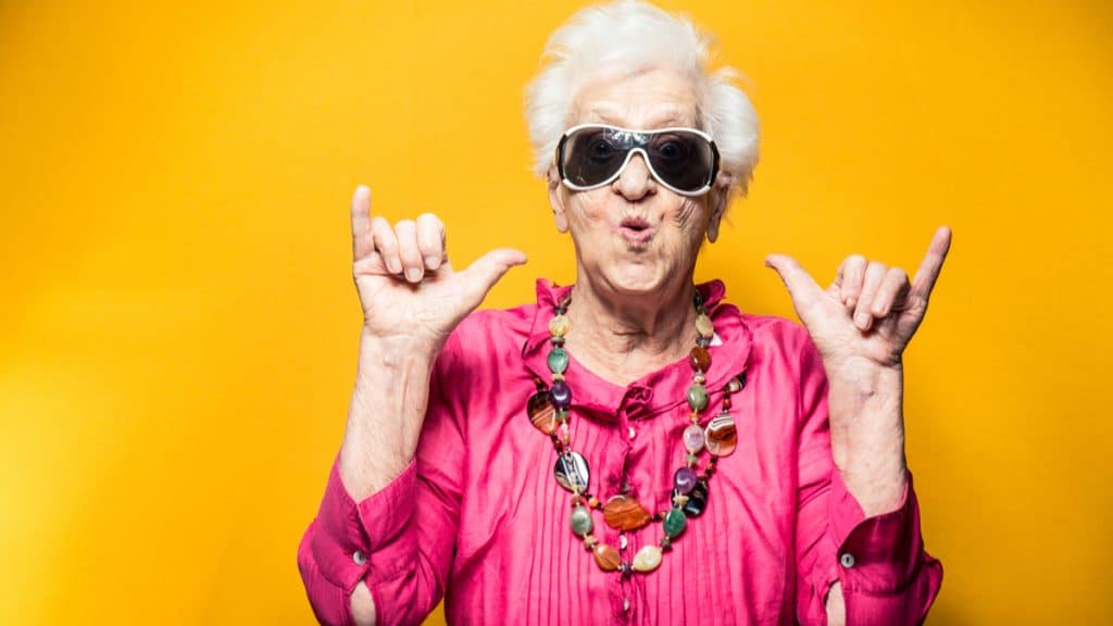 Senhora de idade vestindo trajes coloridos e óculos escuros, fazendo uma pose mais jovial à frente de um fundo alaranjado. A ideia da imagem é passar longevidade e juventude mesmo em idades avançadas