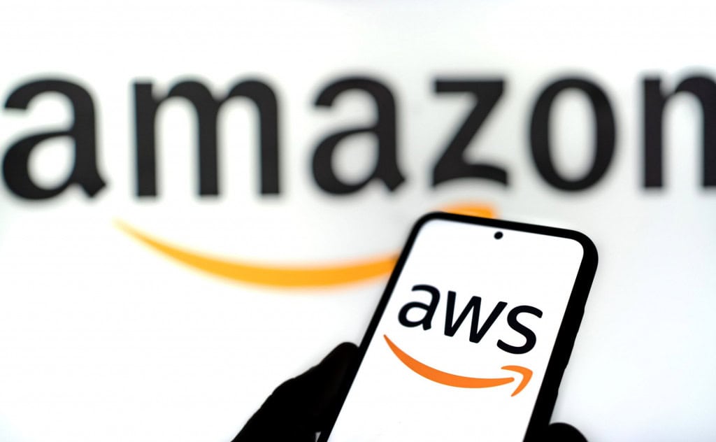 Ao fundo, desfocado, o logo da Amazon; à frente, logo da AWS em um smartphone
