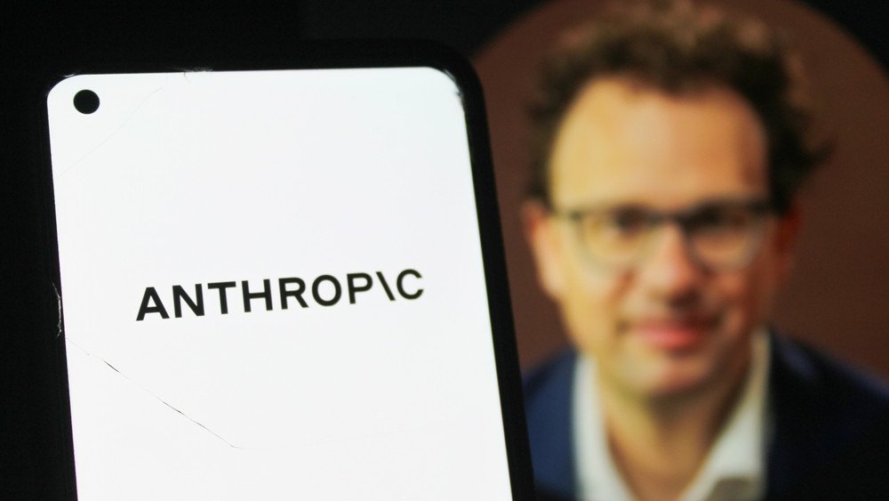 Logo da Anthropic em primeiro plano em um smartphone; ao fundo, imagem desfocada de Dario Amodei