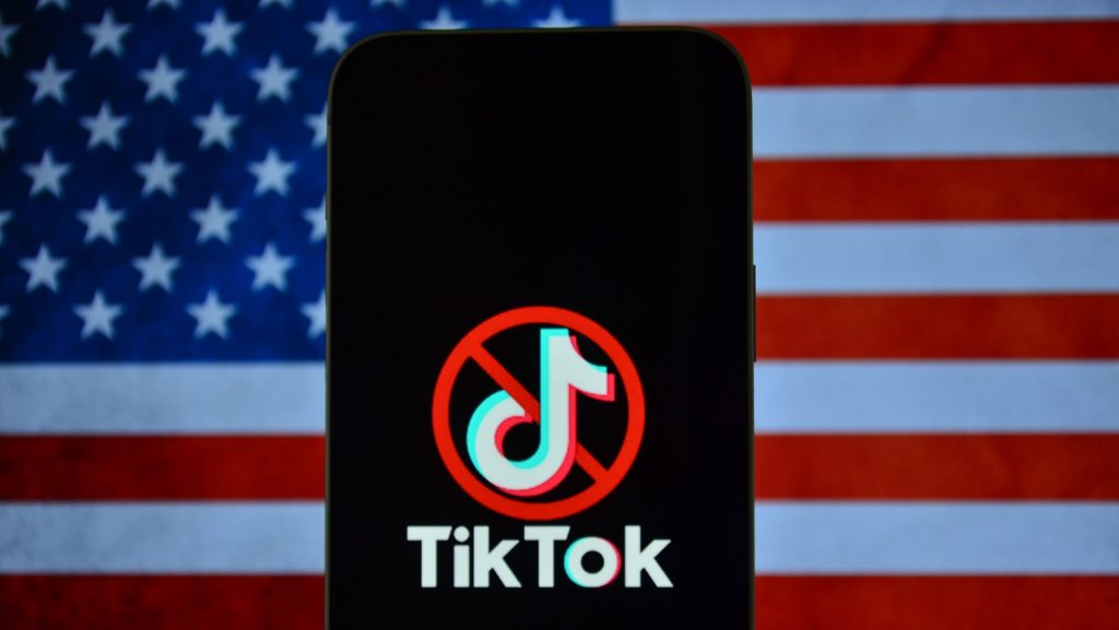 Logo do TikTok com um ícone de proibição por cima em um smartphone; atrás, a bandeira dos Estados Unidos