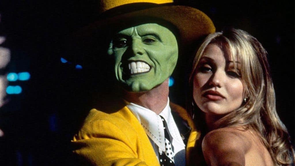 Um homem com o rosto pintado de verde, usando um terno amarelo e chapéu combinando, sorri exageradamente enquanto abraça uma mulher loira de expressão confiante
