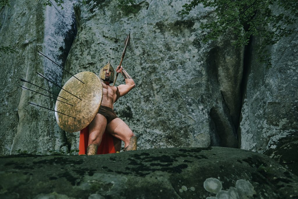 Soldado medieval em pose de ataque, à frente de uma parede de pedra, com a lança na mão esquerda e apontada para a direita, e, na mão direita, um escudo com várias lanças fincadas nele