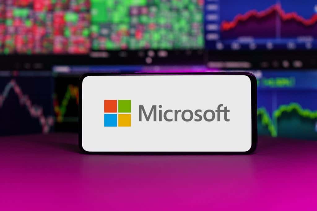 Logo da Microsoft em um smartphone; atrás, telas com gráficos com ações