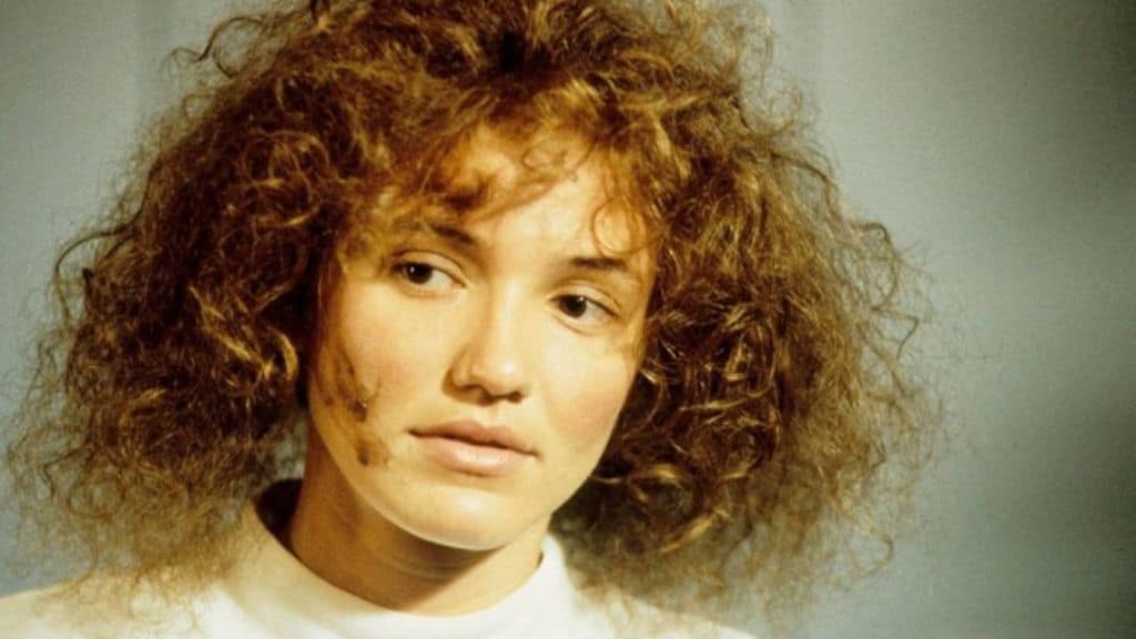 Mulher com cabelo volumoso e encaracolado, usando uma blusa branca, com expressão neutra e uma pequena mancha no rosto